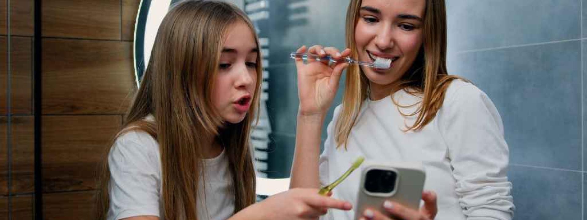 The Truth About Mouthwash, Charcoal Toothpaste, and TikTok Trends: What’s Backed by Science Two young girls smiling, brushing their teeth and looking at a smartphone